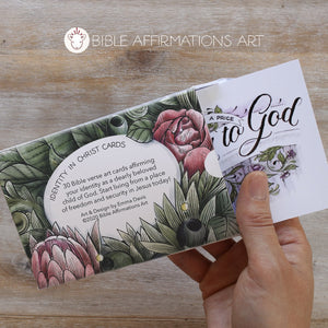 Back of Identity in Christ open card pack with partially protruding floral card emerging from the open box held in a right hand. There are floral illustrations on the box and a blurb that reads "Identity in Christ Cards. 30 Bible verse art cards affirming your identity as a dearly beloved child of God. Start living from a place of freedom and security in Jesus today! Art & Design by Emma Davis copyright 2020 Bible Affirmations Art."