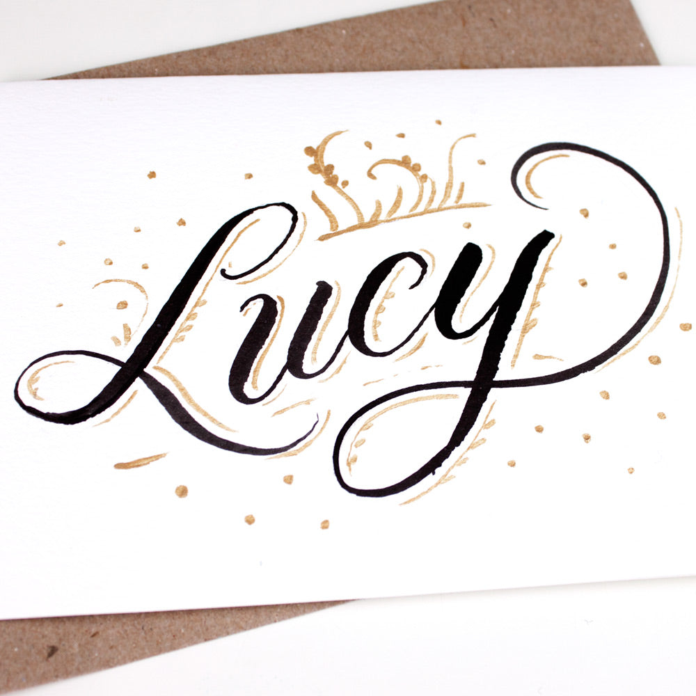 Detail of hand lettered name Lucy in black and gold ink on white card with a kraft envelope
