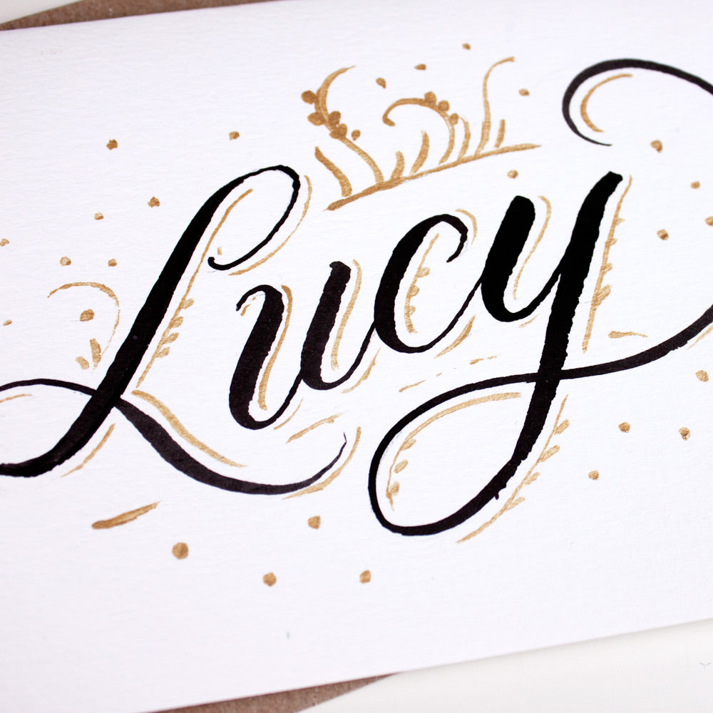 Detail of hand lettered name Lucy in black and gold ink on white card
