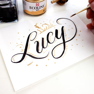 Handwritten card with script "Lucy" handlettered with black and gold ink and hand holding a round watercolor brush