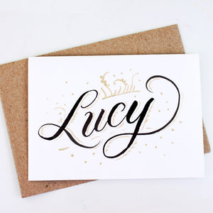 Handwritten card with script "Lucy" handlettered with black and gold ink styled on a kraft envelope
