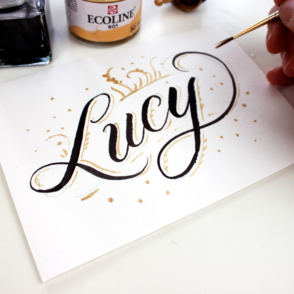 Handwritten card with script "Lucy" handlettered with black and gold ink and hand holding a round watercolor brush