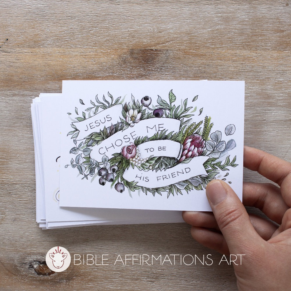 Hand holding postcard with floral illustration with banner reading "Jesus chose me to be his friend." Bible Affirmations Art logo with Lamb icon